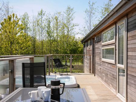 A wooden deck with a glass table and coffee set next to a hot tub and black chairs at 7 Meadow Retreat in Dobwalls