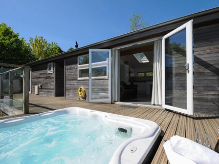 An outdoor hot tub on a wooden deck with glass railing and open double doors leading to a room at 5 Meadow Retreat in Dobwalls