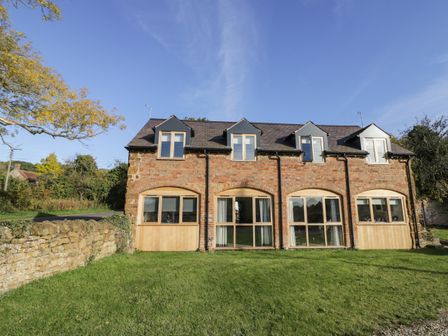 A brick two-story house with dormer windows and large ground floor windows surrounded by a grassy lawn and stone wall at Granary Cottage in Little Dassett near Gaydon