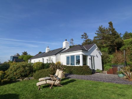 A white single-story house with two chimneys surrounded by bushes and trees with two reclining chairs on a grassy lawn in front at Fron Olau in Nefyn