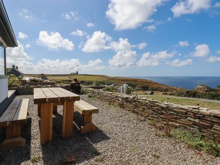 An outdoor area with a wooden picnic table and benches near a stone wall overlooking fields and the sea at Camelot in Tintagel