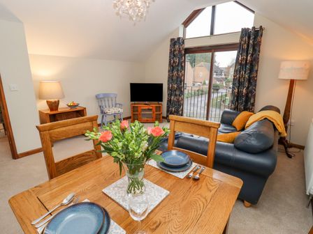 A dining table set with plates and cutlery and a bouquet of flowers next to a living area with a leather sofa a wooden chair a TV and large window at The Coach House in Cheltenham