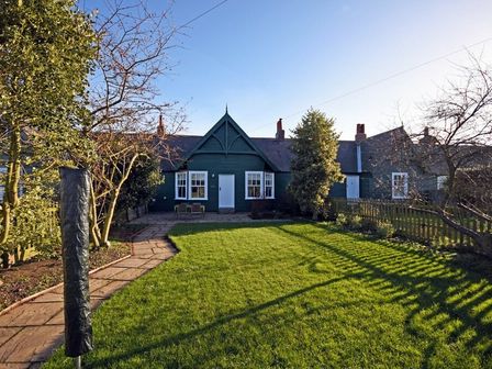 A green lawn with a stone pathway leading to a house with dark green exterior and white doors and windows at Periwinkle in Bamburgh