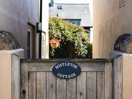 A wooden gate with a sign reading mistletoe cottage between two buildings with a hanging basket of flowers in the background at Mistletoe Cottage in Bamburgh