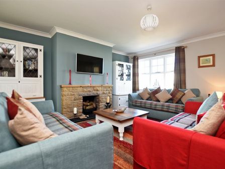 A living room with two light blue sofas a red armchair a wooden coffee table a stone fireplace and a wall-mounted TV at Armstrong Cottages No22 in Bamburgh
