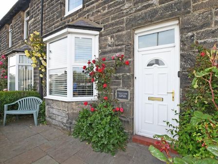 A white front door with a bay window and climbing rose bushes on a stone house with a green bench on the pavement at Cladach Cottage in Seahouses