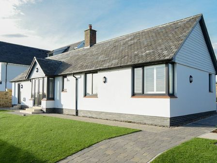 A single story house with white walls and a grey roof with paved path and green lawn at Rock Lodge in Alnmouth