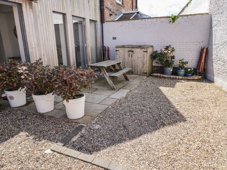 An outdoor patio area with potted plants a wooden picnic bench a storage box and gravel ground at Riverside Lodge in Alnmouth