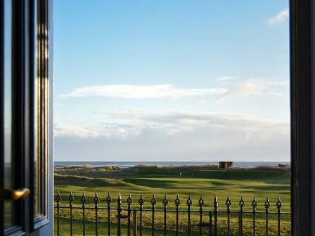 A view of a golf course and the sea through an open doorway with a metal railing outside at Mariners House in Alnmouth