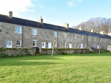 A row of stone cottages with white doors and windows and a grass lawn in front at Farne View Cottage in Seahouses