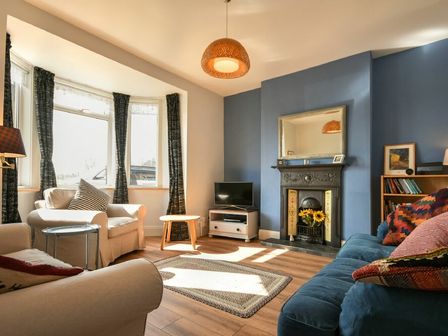 A living room with armchairs, a blue sofa, a TV on a stand, a fireplace with sunflowers, and a bookshelf at Rosedale House in Amble
