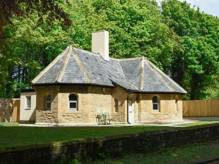 A stone cottage with a slate roof surrounded by trees and a wooden fence at The Lodge (Alnwick) in Alnwick
