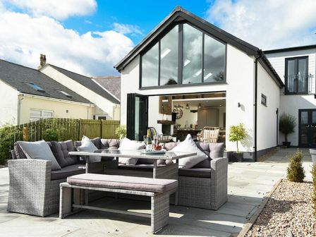 An outdoor patio with grey wicker furniture and cushions outside a white house with large windows at Marmalade Cottage in Seahouses