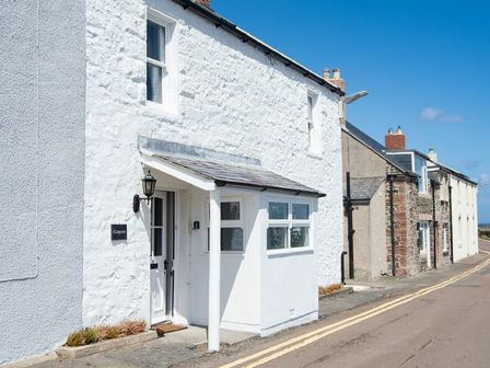 A white stone house with a porch and a lamp on a street with adjacent houses at Cargoes in Craster
