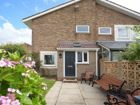 The front exterior of a brick house with a black door two wooden benches and a paved path and flowering plants at Coastal Plaice in Beadnell