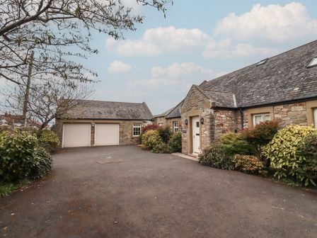 An outdoor driveway with a stone building featuring two garage doors and a white entrance door surrounded by bushes at The Wynd in Bamburgh