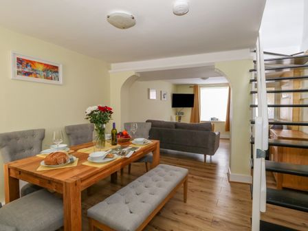 A dining area with a wooden table, chairs, a bench, and table settings next to a living room with a sofa and a wall-mounted TV at Cove Street Cottage in Weymouth