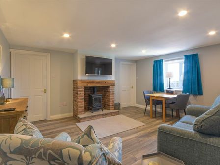 A living room with a brick fireplace TV sofa armchair wooden table and blue curtains at Dairymaid's Cottage in Amble