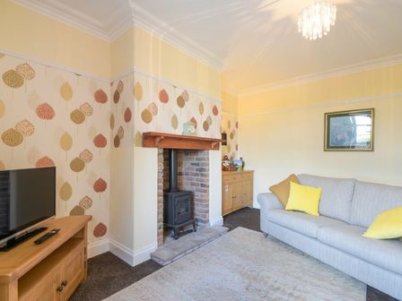 A living room with a television on a wooden stand a sofa with yellow cushions a fireplace with a wood burning stove and a wooden cabinet at Dairyman's Cottage in Amble