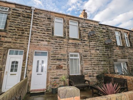 Stone front facade of a terraced house with white doors and windows a wooden bench and potted plants in the front yard at Creel Cottage Blackwood Street in Amble