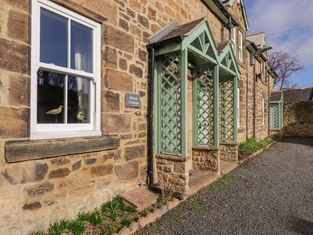 The exterior of a stone cottage with green lattice porch structures and a window with two bird figurines at Willow Cottage Lesbury