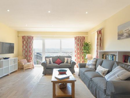 A living room with two gray sofas a wooden coffee table a wicker chair a flat screen television and large windows with patterned curtains at The Boathouse Alnmouth