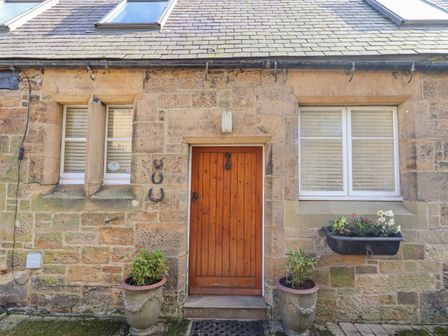 A stone exterior wall with a wooden door between two windows and flower pots in front at Coach House (Alnmouth) in Alnmouth