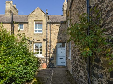 The exterior of a stone cottage with a white door and windows surrounded by bushes and plants at Avocet Cottage in Bamburgh