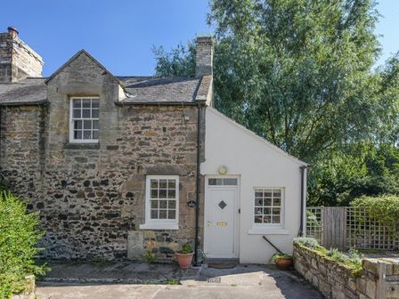 A stone and white-painted cottage exterior with windows and a door surrounded by trees and plants at Dunlin Cottage in Bamburgh