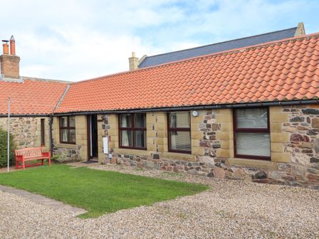 A stone cottage with red roof tiles a red bench on the grass outside at Kittling Cottage in Bamburgh