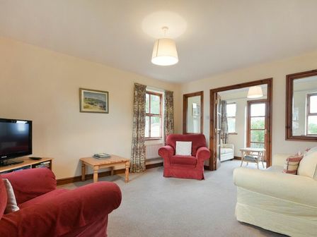 A living room with red armchairs a cream sofa a wooden table a television and windows at Smithy Court in Craster