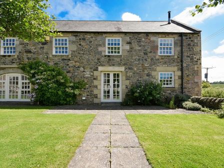 Stone house exterior with tiled pathway leading to white-framed glass door and windows surrounded by green lawn and bushes at Budle Sands in Bamburgh