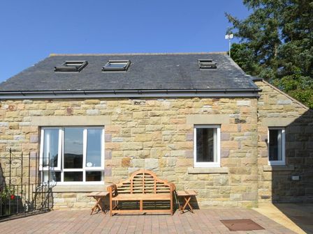 Exterior of a stone cottage with three windows and a wooden bench on a paved patio at Mossyford Cottage in Alnwick