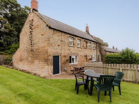 A stone cottage with black door and windows with garden furniture and a wooden fence at Bilton Farm Cottage No1 in Alnmouth