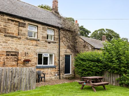 A stone cottage with closed windows a black door a wooden picnic table green bushes and a lawn at Bilton Farm Cottage No2 in Alnmouth