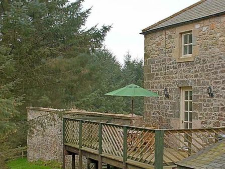 A stone house with a wooden balcony and a green umbrella surrounded by trees at The Bolthole in Eglingham