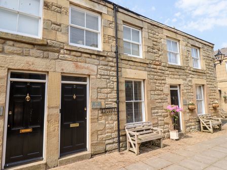 A stone building with black doors and windows with blinds and wooden benches outside at Aln Cottage in Alnwick