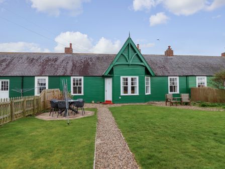 A green cottage with a gravel path leading to the door and outdoor seating areas at Armstrong Cottages No2 in Bamburgh