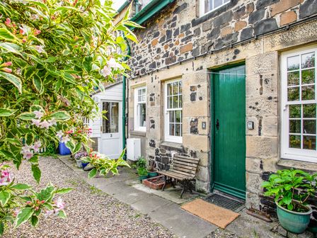 Stone cottage exterior with green door wooden bench potted plants and gravel pathway at Elliot Cottage in Bamburgh