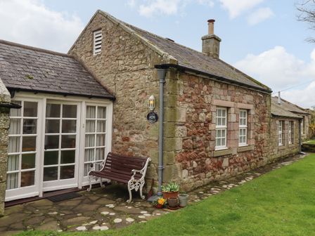 An exterior view of a stone cottage with white framed windows a bench and potted plants near the door at Glebe Cottage in Bamburgh