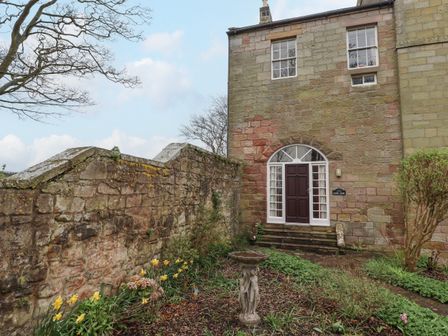 A stone house with a dark brown door and white windows next to a stone wall and garden with plants and flowers at Glebe House in Bamburgh