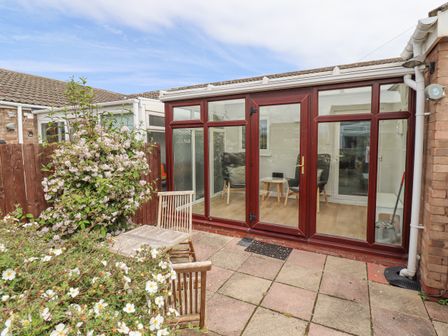 A patio with wooden chairs and table next to flowering plants and glass doors leading to a room at Bedwine Cottage in Beadnell