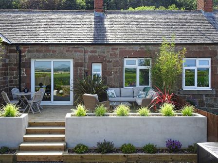 An outdoor patio with wooden table and chairs a cushioned seating area and plants in planters at Bellshill Cottage in Belford
