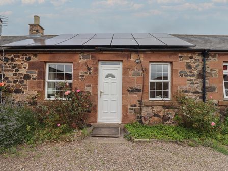 Stone cottage exterior with white door and two windows with solar panels on roof at Budle Bay Cottage in Bamburgh