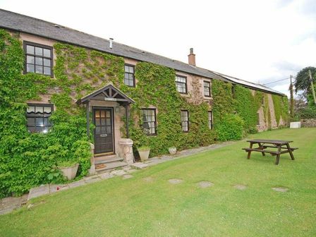 The exterior of a stone cottage with ivy growing on the walls and a wooden picnic table on a grassy lawn at Croft Cottage (Yeavering) in Wooler