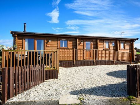 A wooden cabin with a fenced gravel driveway under a blue sky at Heather Cottages - Brown Owl in Bamburgh