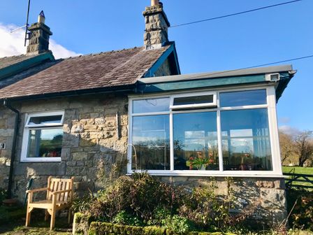 A stone house with a wooden chair outside and a large window showing flowers inside at Leazes Cottage in Rothbury
