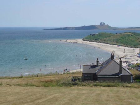 A coastal view with a house in the foreground a beach with people and a castle on a hill across the water at Melvin Cottage in Low Newton-By-The-Sea