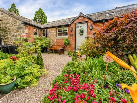 A garden with flowers and plants in front of a brick and stone house with a bench and a door with a wreath at Nurses Cottage in Alnwick