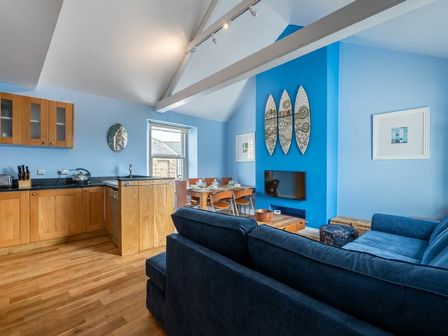 A living area with blue walls a wooden kitchen open to a dining table and blue sofa at Pilots Rest in Alnmouth